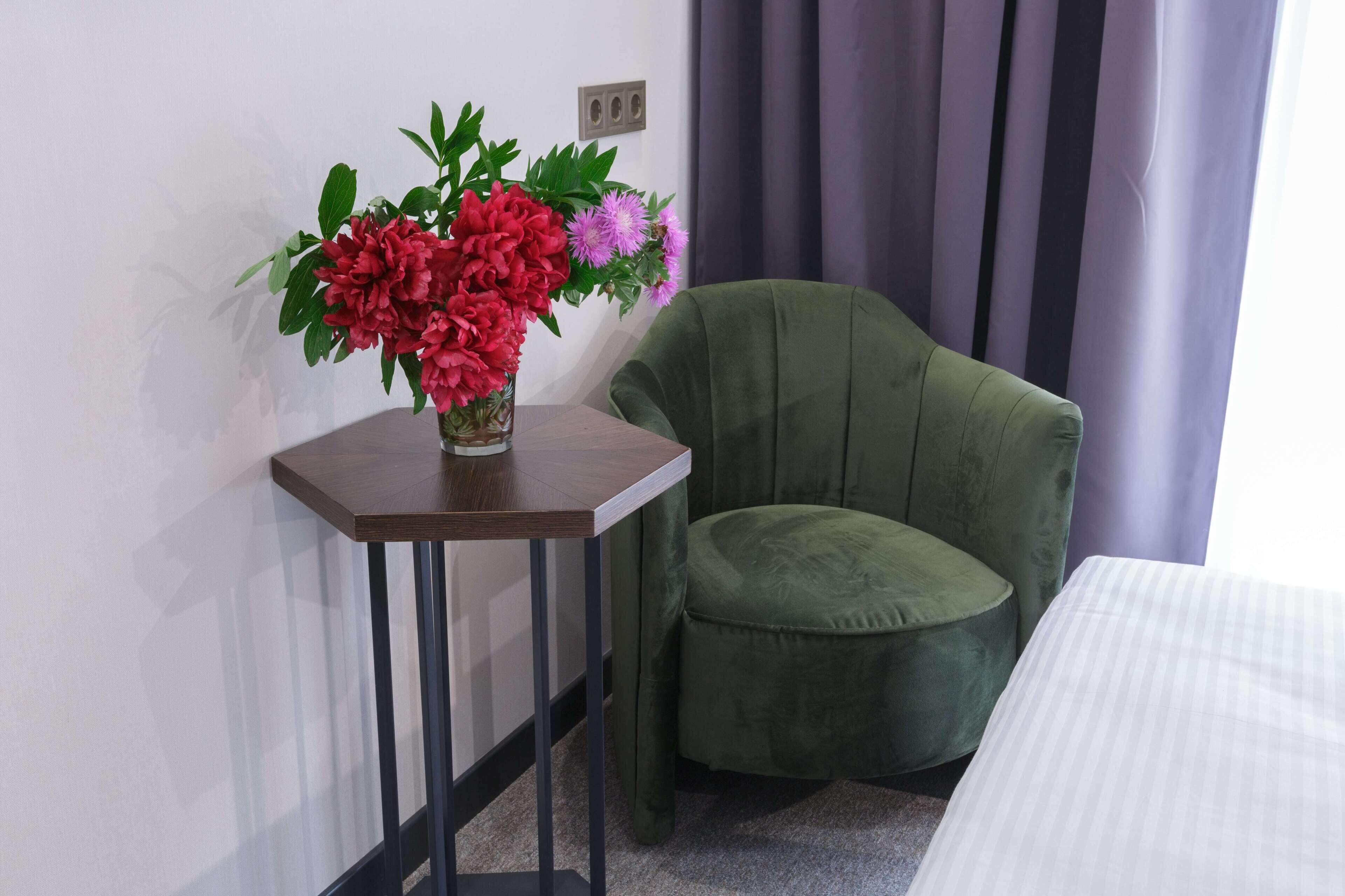 A room with a green upholstered chair, a small wooden table with a vase of flowers, and a bed with white bedding.