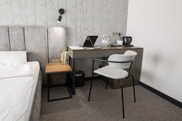 Habitación de hotel con cama, escritorio con una computadora portátil, una silla, un cesto de basura y una cafetera.