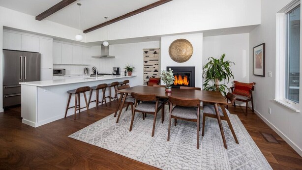 A modern kitchen with white cabinets, a central island with bar stools, a fireplace, and a dining area with a wooden table and chairs. - Hawks Perch - New Downtown Modern, Panoramic View (Hood River)