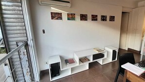Ein Zimmer mit weißer Wand, einer kleinen weißen Schrankwand mit Büchern, einem schwarzen Stuhl und einem Schreibtisch mit Laptop und Papieren.