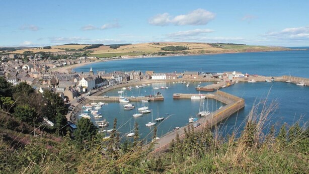 Une ville côtière dotée d’un port de plaisance, de bâtiments et d’un paysage vallonné en arrière-plan.