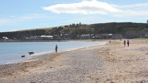 Une plage de galets bordée d’une mer calme, quelques personnes qui se promènent et des maisons en arrière-plan.