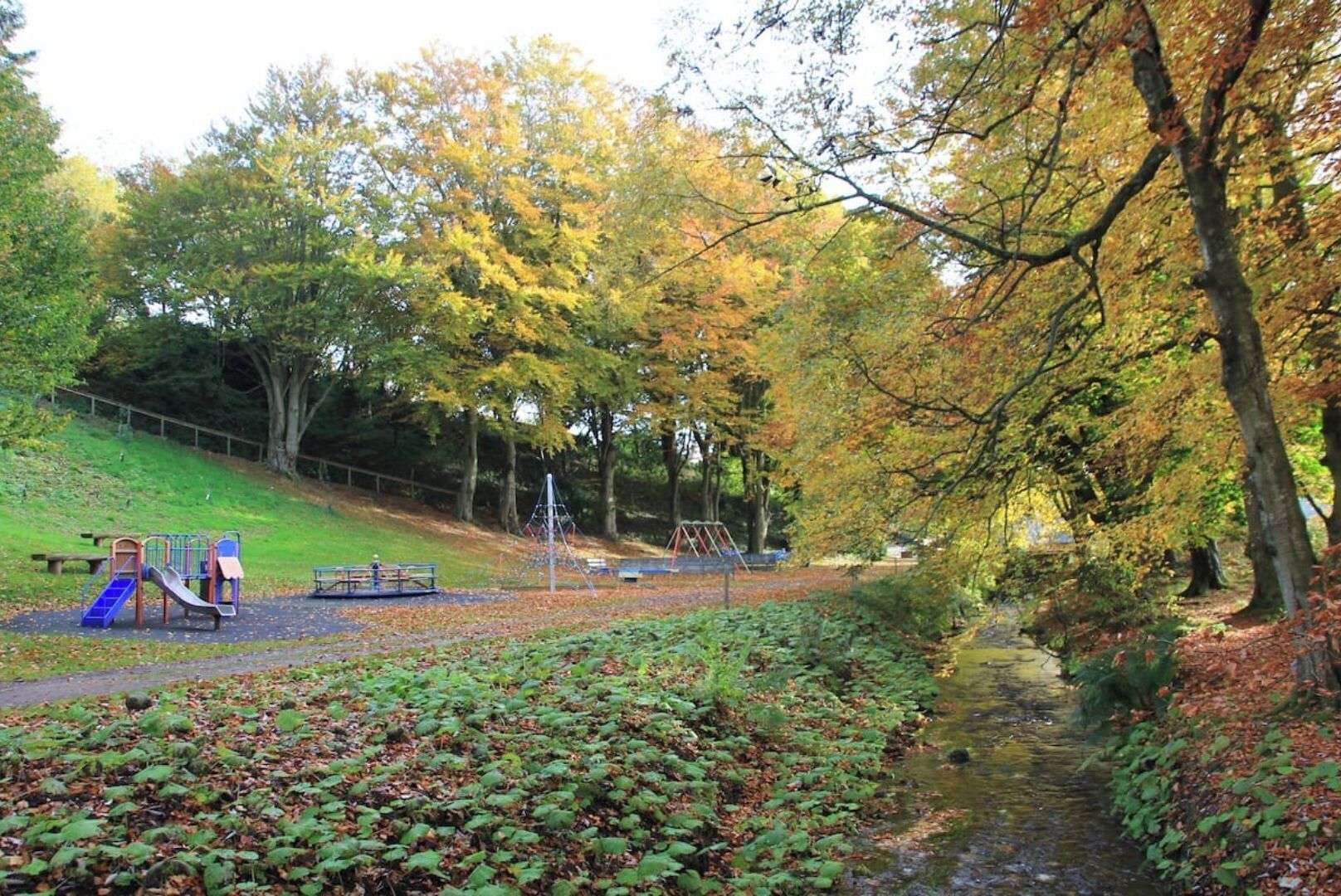 A park with playground equipment, a grassy slope, and a stream.