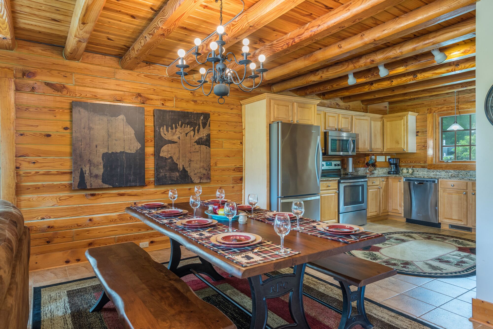 Interior de una cabaña de madera con una mesa de comedor para seis personas, una araña y una cocina con electrodomésticos modernos.