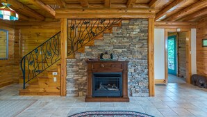 L’intérieur d’une cabane en rondins, avec une cheminée en pierre, un escalier en bois et une vue sur l’extérieur.