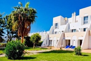 A modern building with white walls and multiple balconies, surrounded by greenery and palm trees.