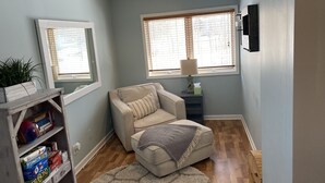 Ein Zimmer mit einem beigefarbenen Sessel, einem kleinen Tisch mit Lampe, einem Bücherregal und einem großen Fenster mit Jalousien.