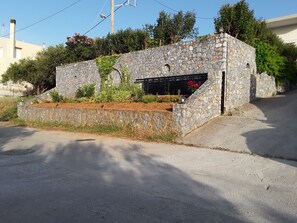 A stone structure with a small garden area, a metal gate, and a paved road in the foreground. - Marianos Apartments (Kissamos)