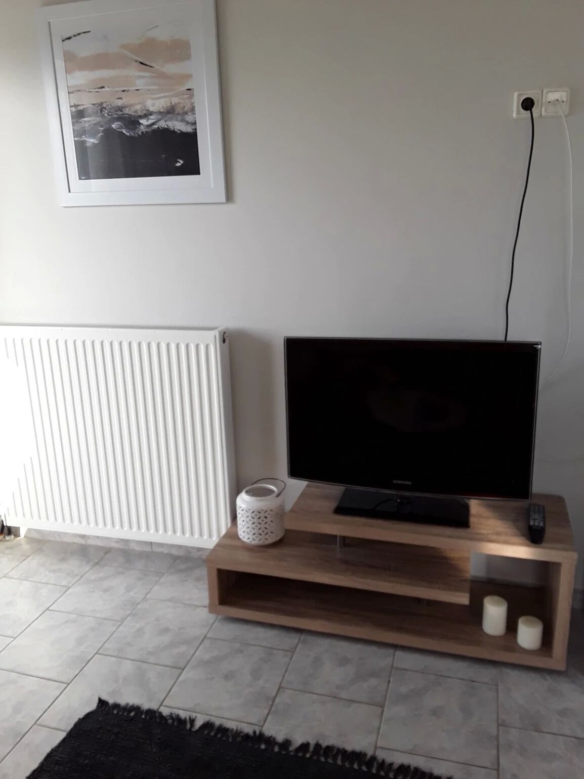 Uma sala de estar com uma televisão de tela plana sobre um suporte de madeira, um radiador branco e um quadro na parede.