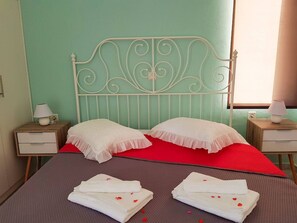 A neatly made bed with white pillows and a red bedspread, two bedside tables with lamps, and a window with curtains. - Marianos Apartments (Kissamos)