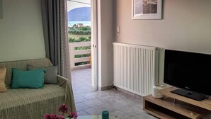 A living room with a sofa, a TV, a table with flowers, and a view of the outdoors. - Marianos Apartments (Kissamos)