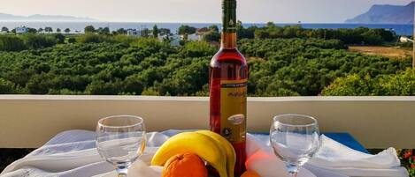 A table with a bottle of wine, two glasses, and fruits on a balcony with a scenic view.