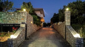 A stone pathway leading to a modern house with a balcony, surrounded by greenery. - Marianos Apartments (Kissamos)