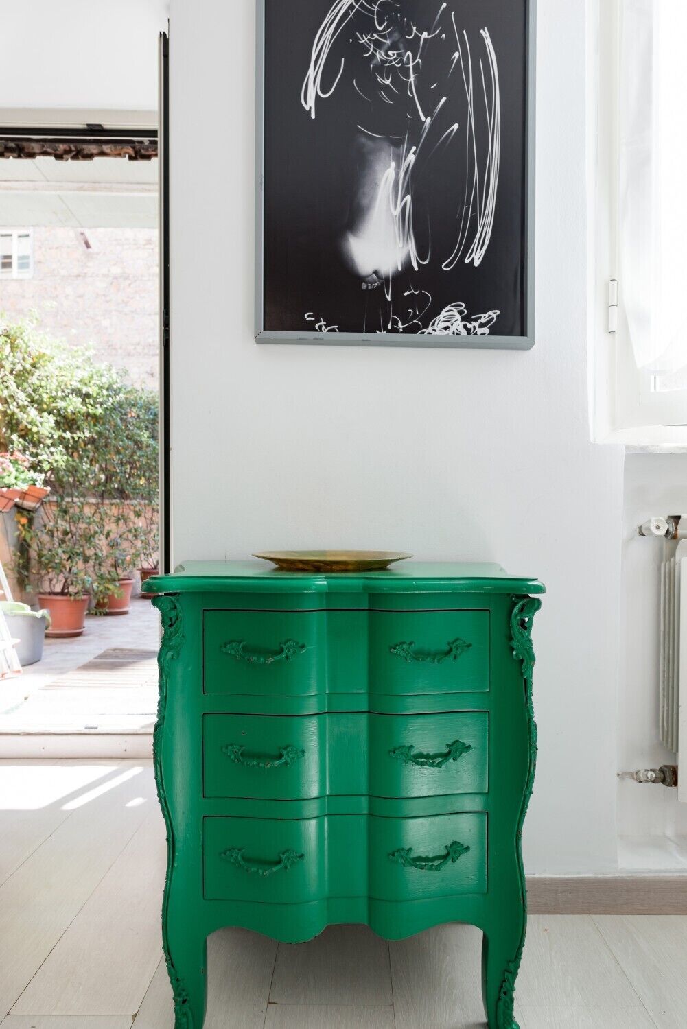 Une commode verte avec une tablette sur le dessus, une peinture abstraite en noir et blanc au-dessus, et des plantes en pot devant une fenêtre.