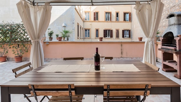 Une table en bois, sur laquelle sont posés une bouteille de vin et un verre, est placée sous une sorte d’auvent agrémenté de rideaux, d’où l’on aperçoit un bâtiment doté de nombreuses fenêtres et d’un four en pierre.