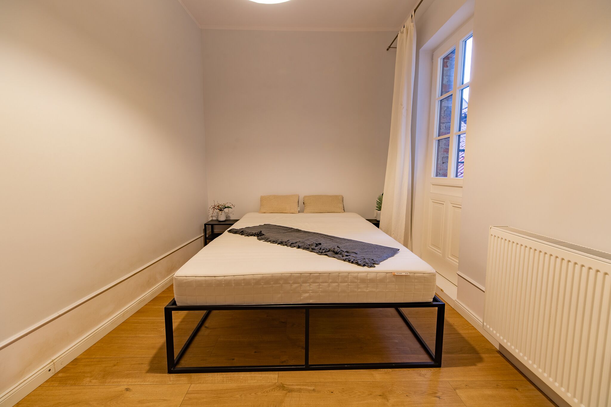 A narrow room with a bed, a nightstand, a window, and a radiator.