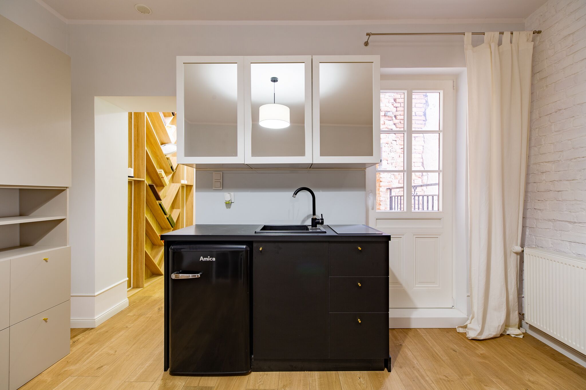 A compact kitchen with white cabinets, a black refrigerator, and a window with white curtains.