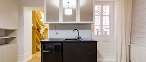 A compact kitchen with white cabinets, a black refrigerator, and a window with white curtains.