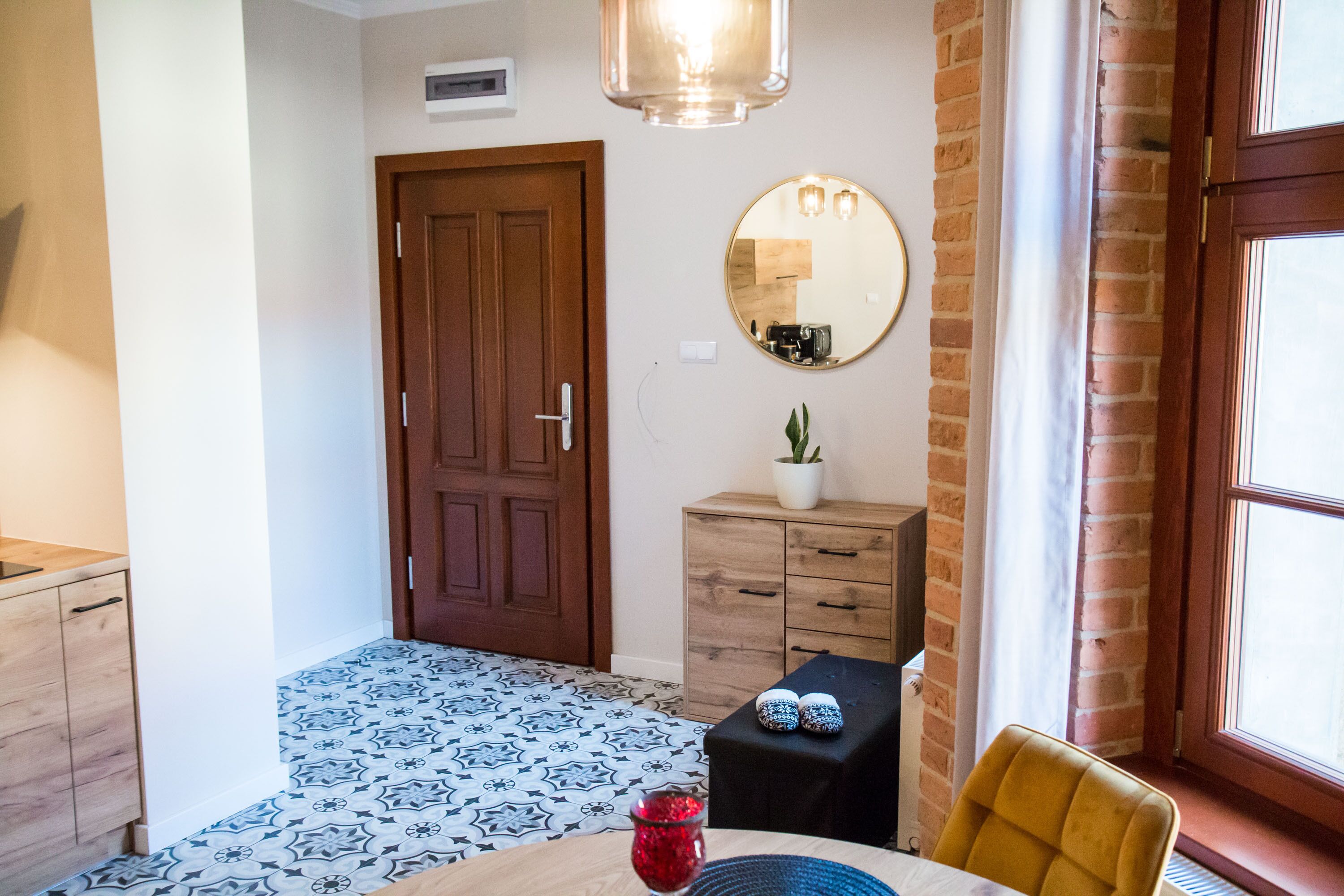 Een kamer met een houten deur, een spiegel, een klein tafeltje met een rode vaas en een gele stoel.