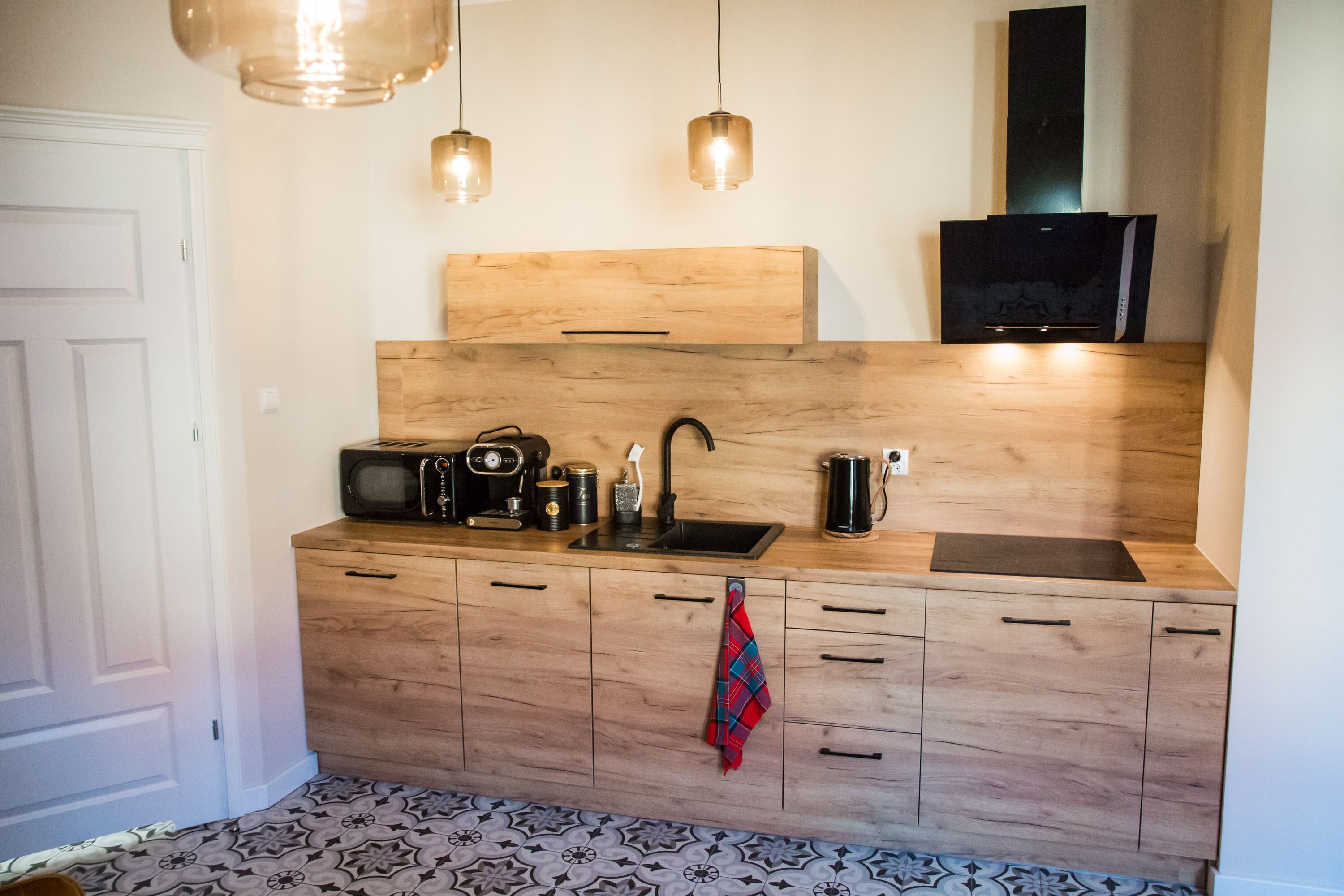 A compact kitchen with a wooden countertop, a microwave, a kettle, and a sink. There are hanging pendant lights and a red towel hanging on the cabinet.