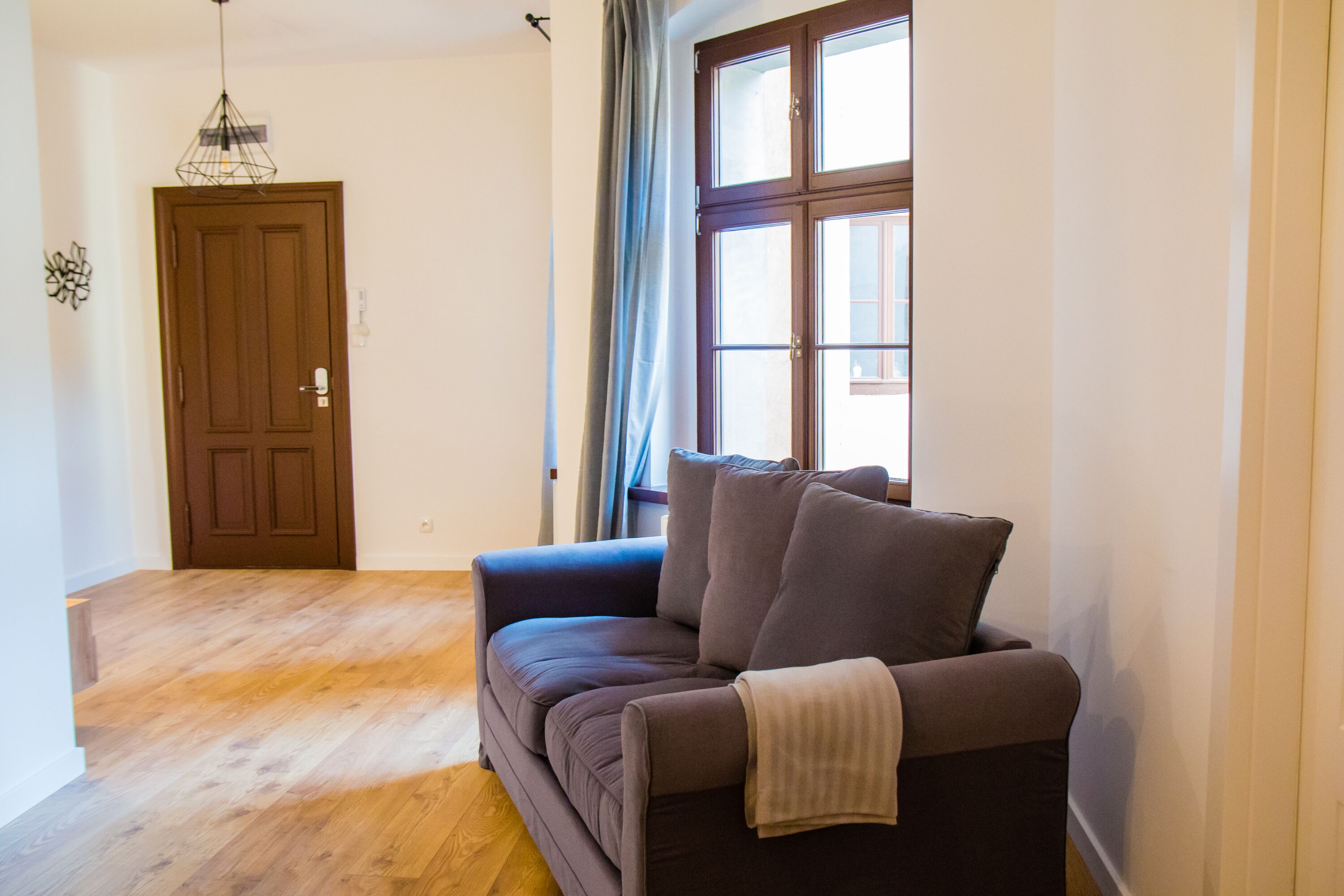 Un salon avec un canapé, un parquet, une porte et une fenêtre avec des rideaux.