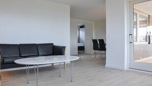 Un salon moderne avec un canapé en cuir noir, une table ronde blanche aux pieds en métal et une table basse en verre. Une porte donne accès à une chambre à coucher équipée d’un lit et d’une chaise. La pièce est dotée d’un parquet en bois clair et de murs blancs.