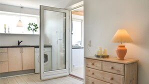 Une cuisine moderne avec une porte vitrée donnant sur un balcon, des meubles de rangement en bois, un lave-linge et une commode en bois surmontée d’une lampe.
