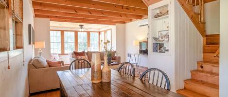 Une table à manger en bois avec des chaises, un lustre, un escalier et un coin salon confortable avec un canapé et un téléviseur.