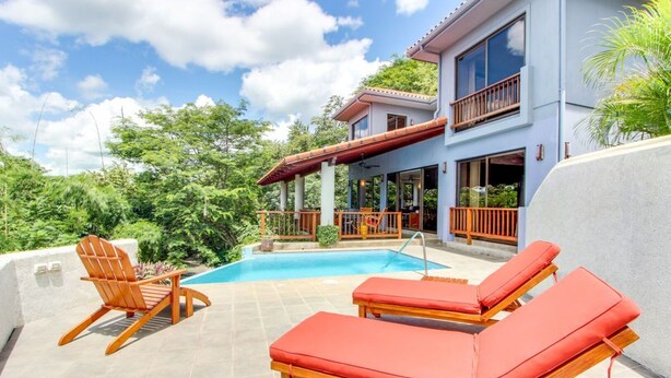 Una casa moderna de dos pisos con piscina, terraza de madera y mobiliario exterior.