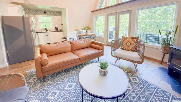 A spacious living room with a sofa, armchair, and coffee table, a kitchen in the background, and large windows offering a view of the outdoors. - Hidden A-Frame at Double Bridge with Firepit (Mountain View)