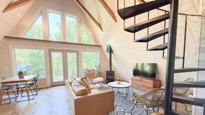 A loft-style room with a wooden floor, a sofa, a TV, and a staircase. - Hidden A-Frame at Double Bridge with Firepit (Mountain View)