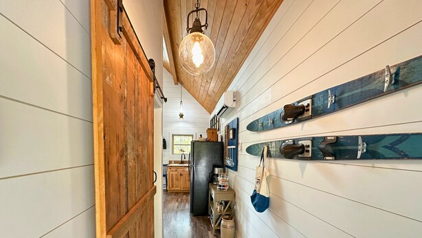 Un pasillo estrecho con paredes y puerta de madera, que conduce a una zona de cocina con un refrigerador y varios utensilios.