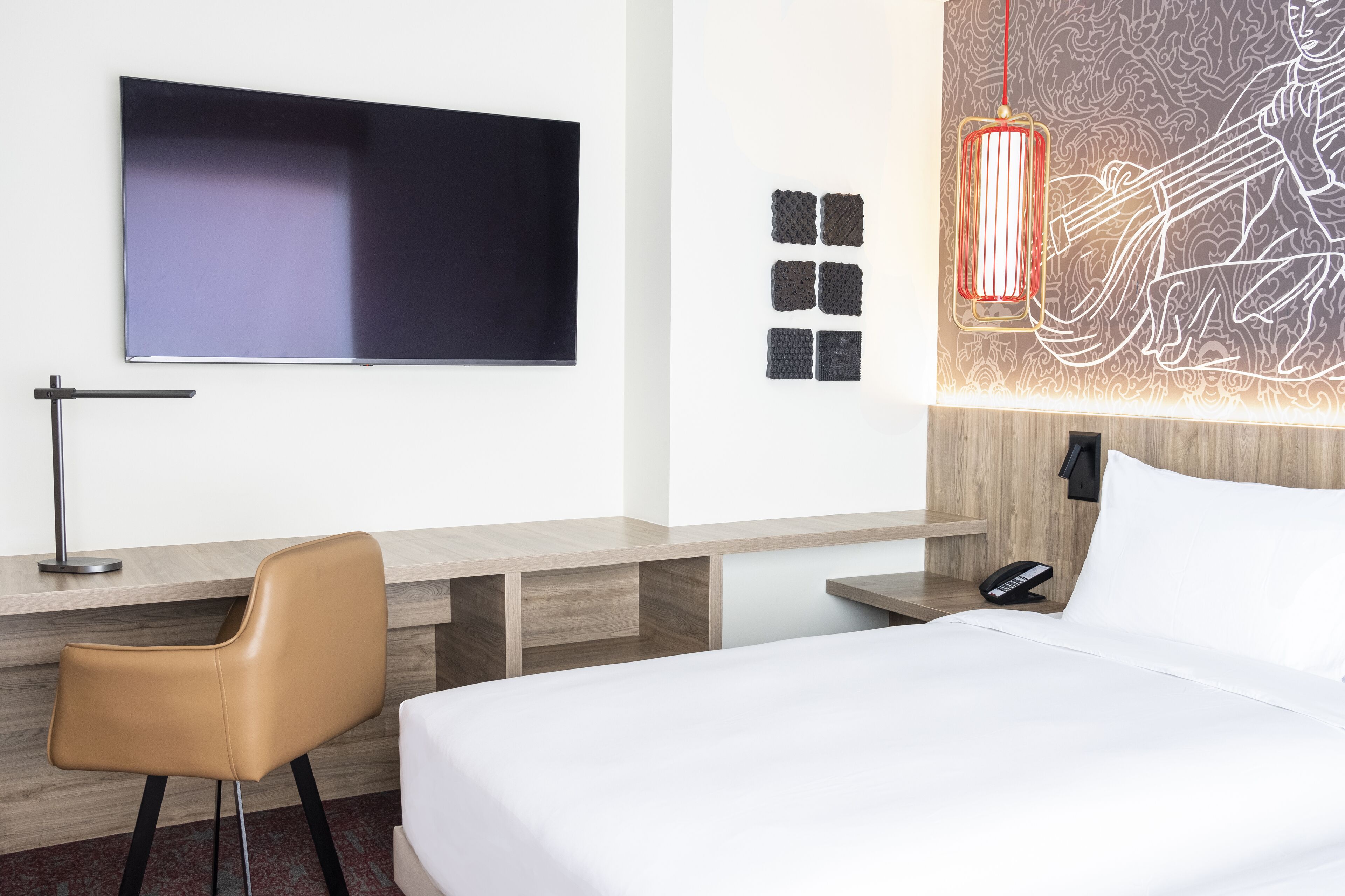 Habitación de hotel con cama, escritorio con silla, televisor de pantalla plana, obra de arte en la pared y lámpara colgante roja.