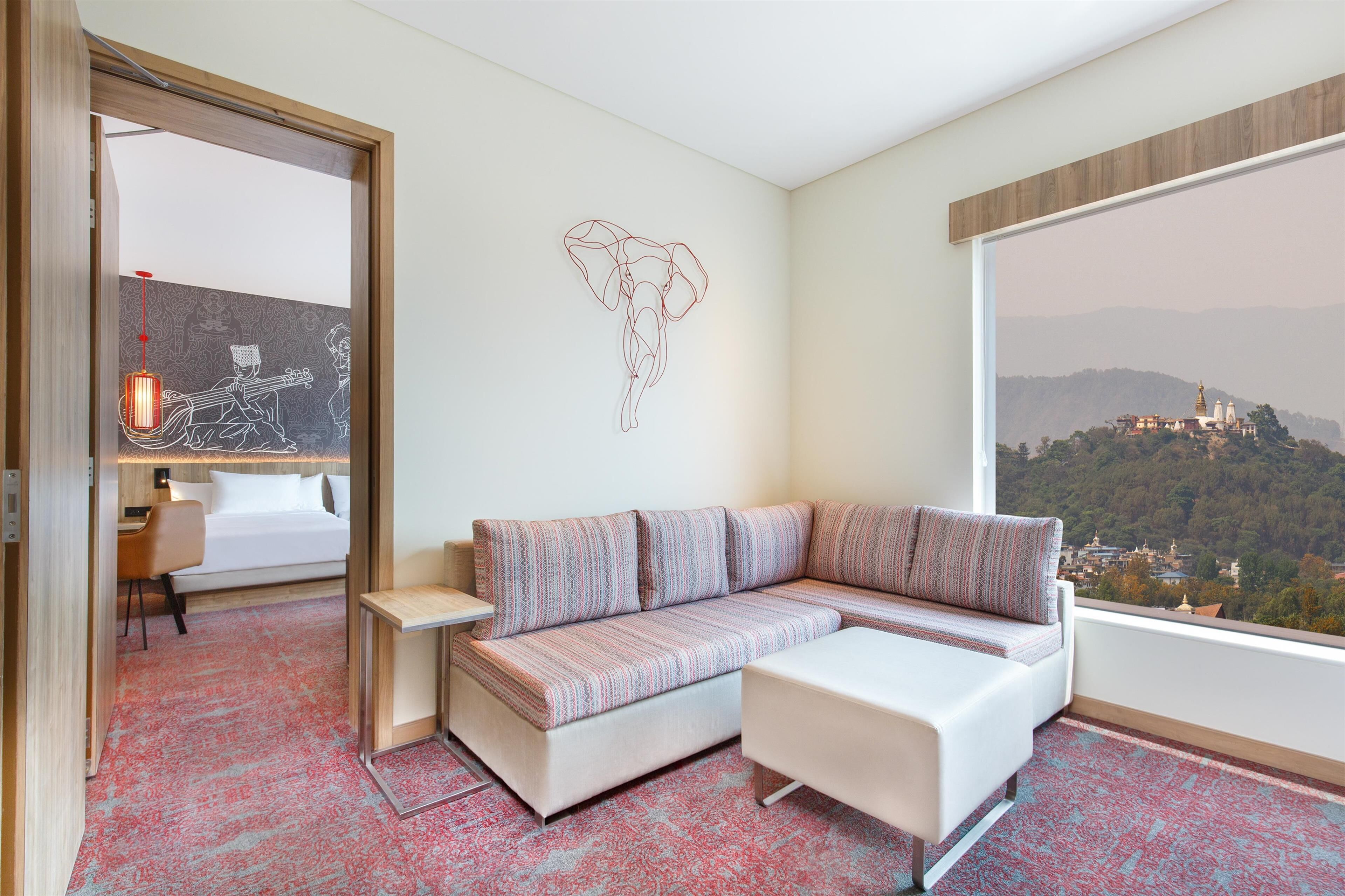 Habitación de hotel moderna con sofá, una mesita, una cama y un amplio ventanal con vistas panorámicas de una colina con edificios.