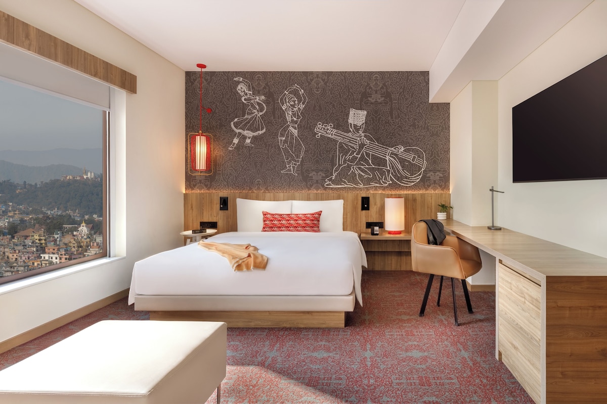 A modern hotel room with a large bed, a desk, a chair, a TV, and a city view.