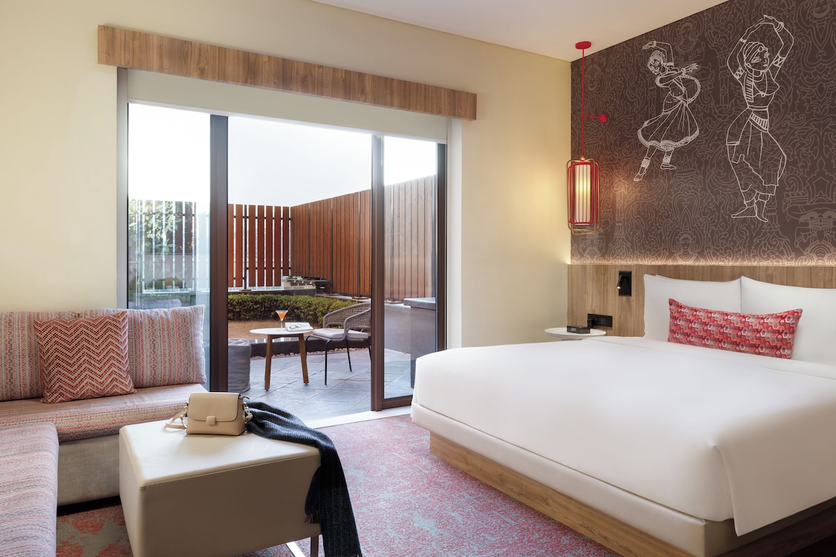 A modern hotel room with a large bed, a sofa, a small table, and a view of the outdoor patio.