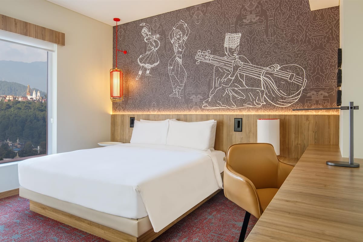 A hotel room with a large bed, a desk, a chair, a wall mural, and a view of a cityscape.