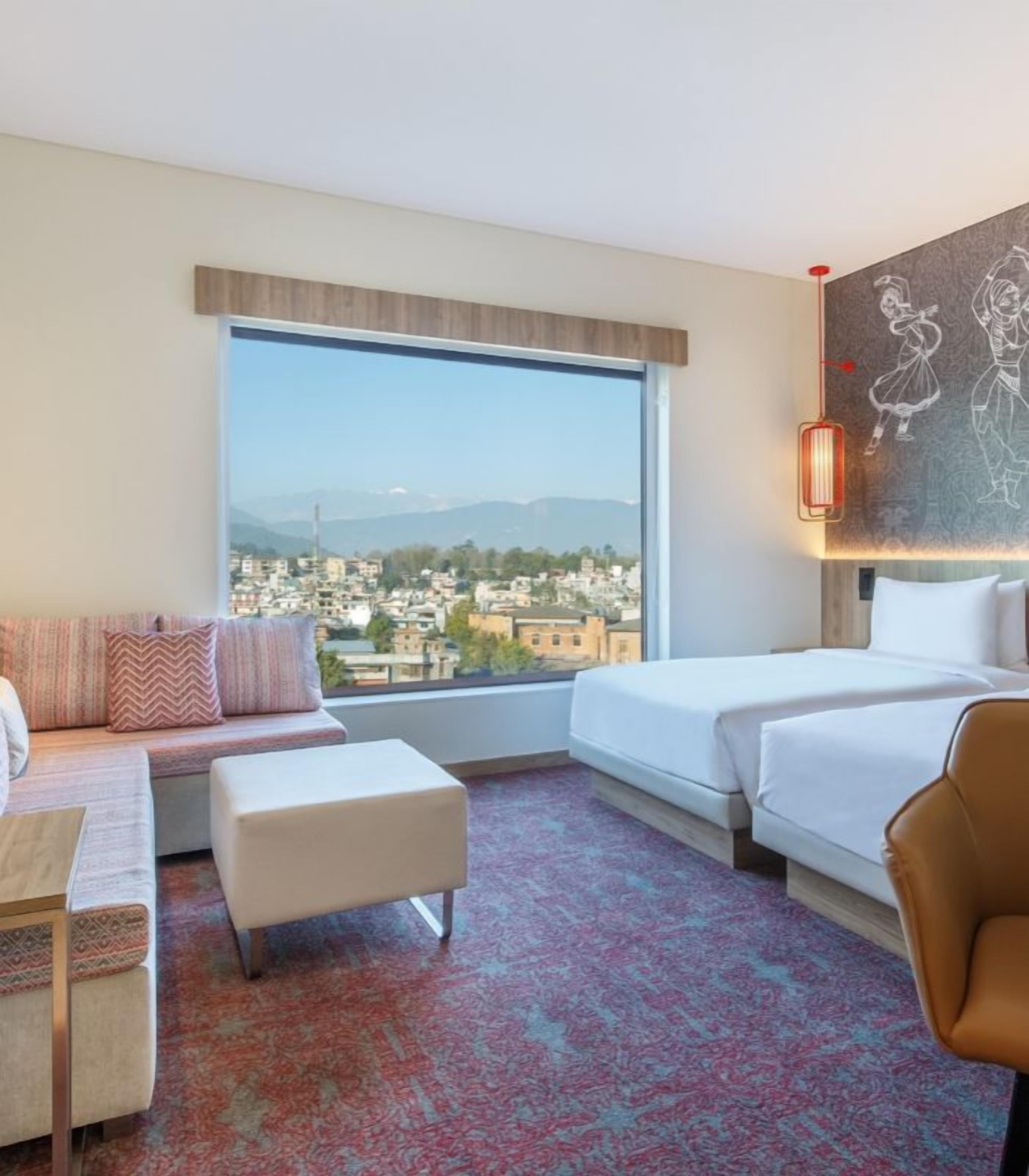 A hotel room with a large window, a sofa, a bed, a desk, and a chair.