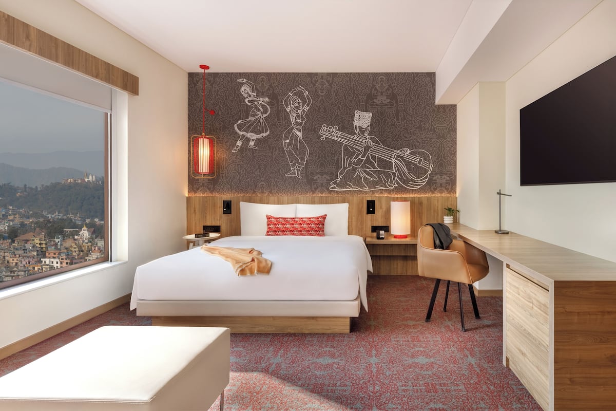 A modern hotel room with a large bed, a desk, a chair, a TV, and a city view.