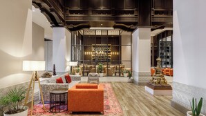 Un vestíbulo amplio con suelo de madera, una zona de estar central con sofás naranjas, una mesa de centro y un tapete decorativo. Hay plantas en macetas y una lámpara grande.