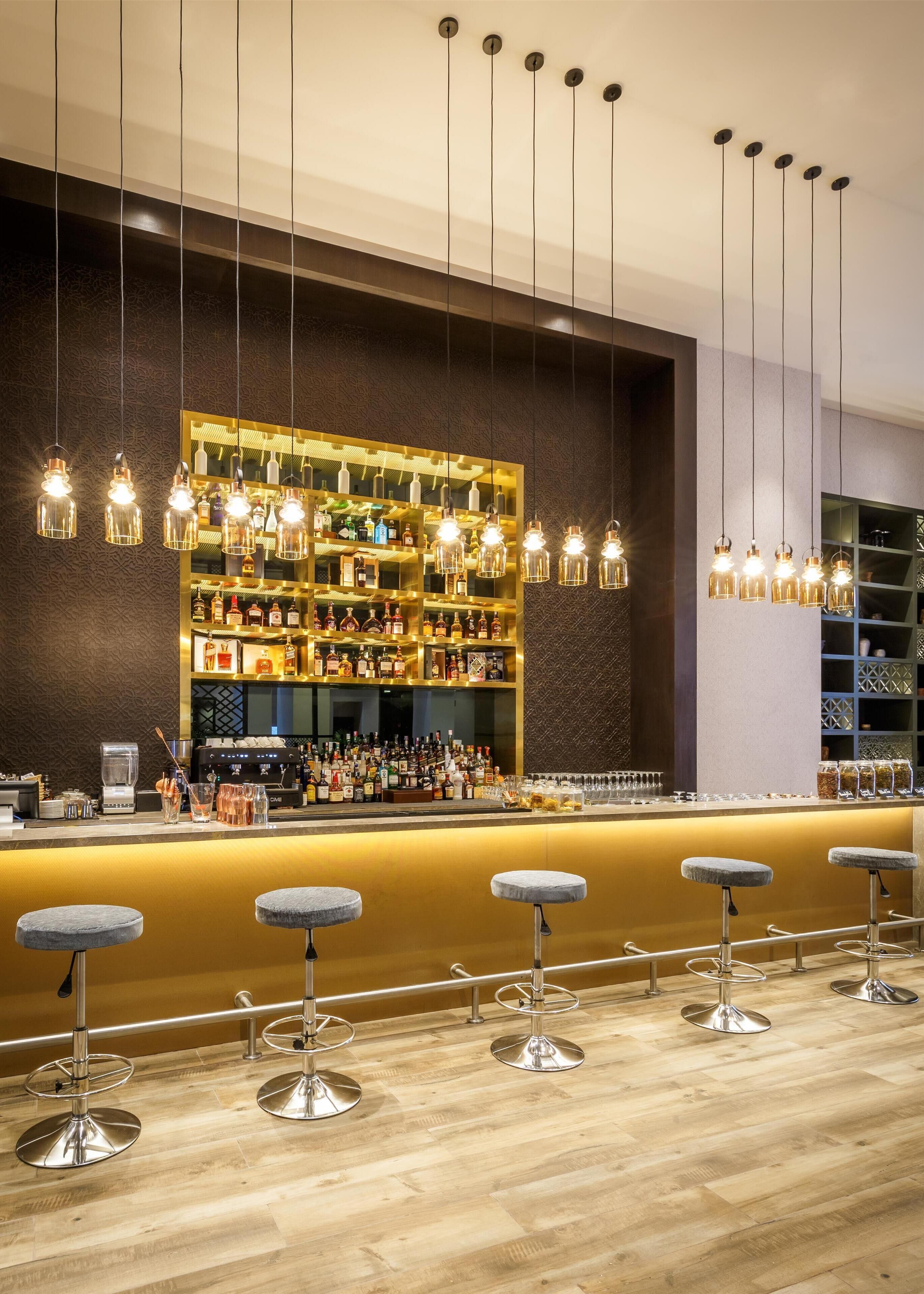 A bar area with a wooden floor, bar stools, and a well-stocked shelf with various bottles.
