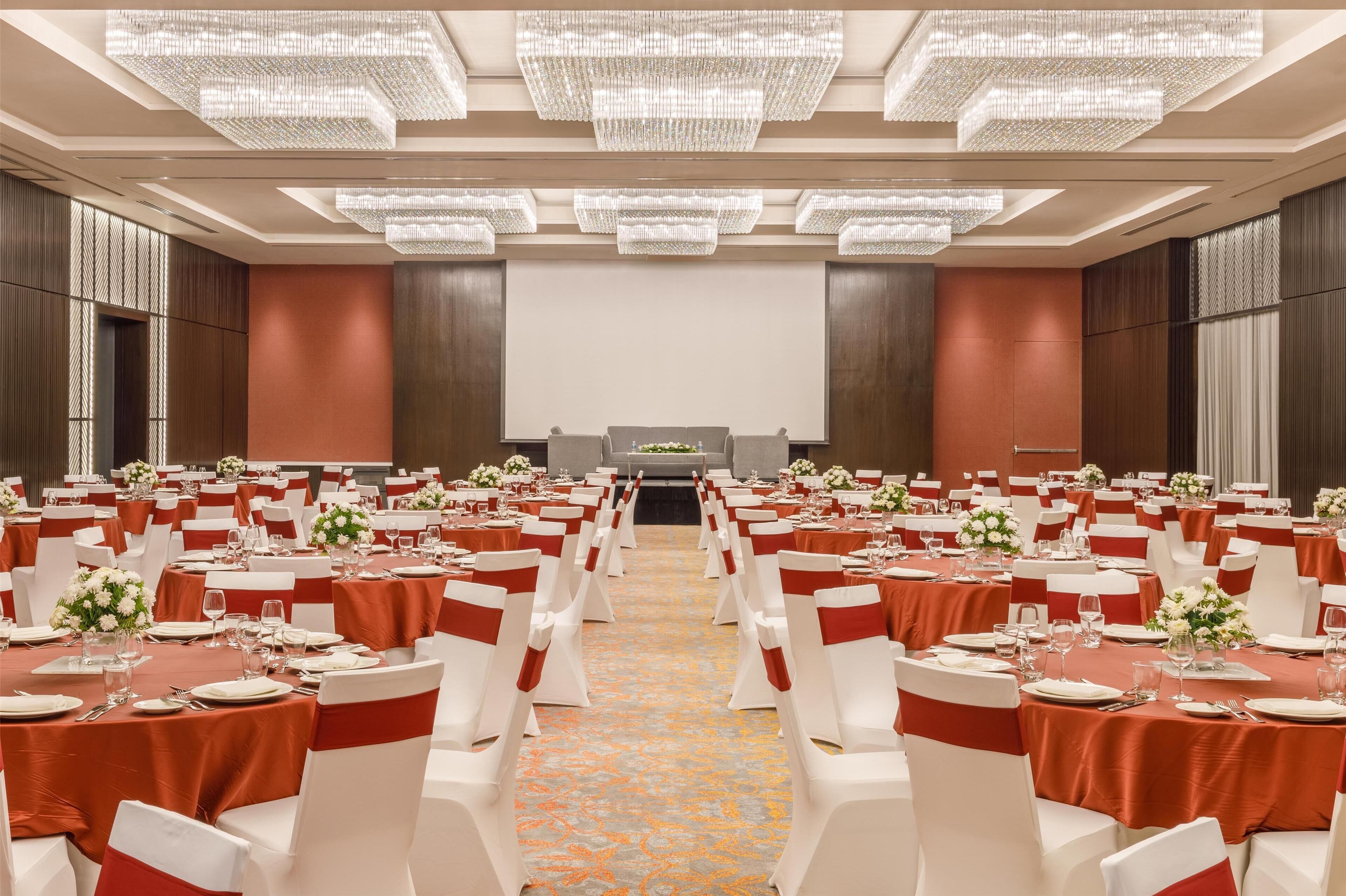 Un salón de banquetes con filas de mesas, sillas blancas con cintas rojas y una pantalla grande al frente.