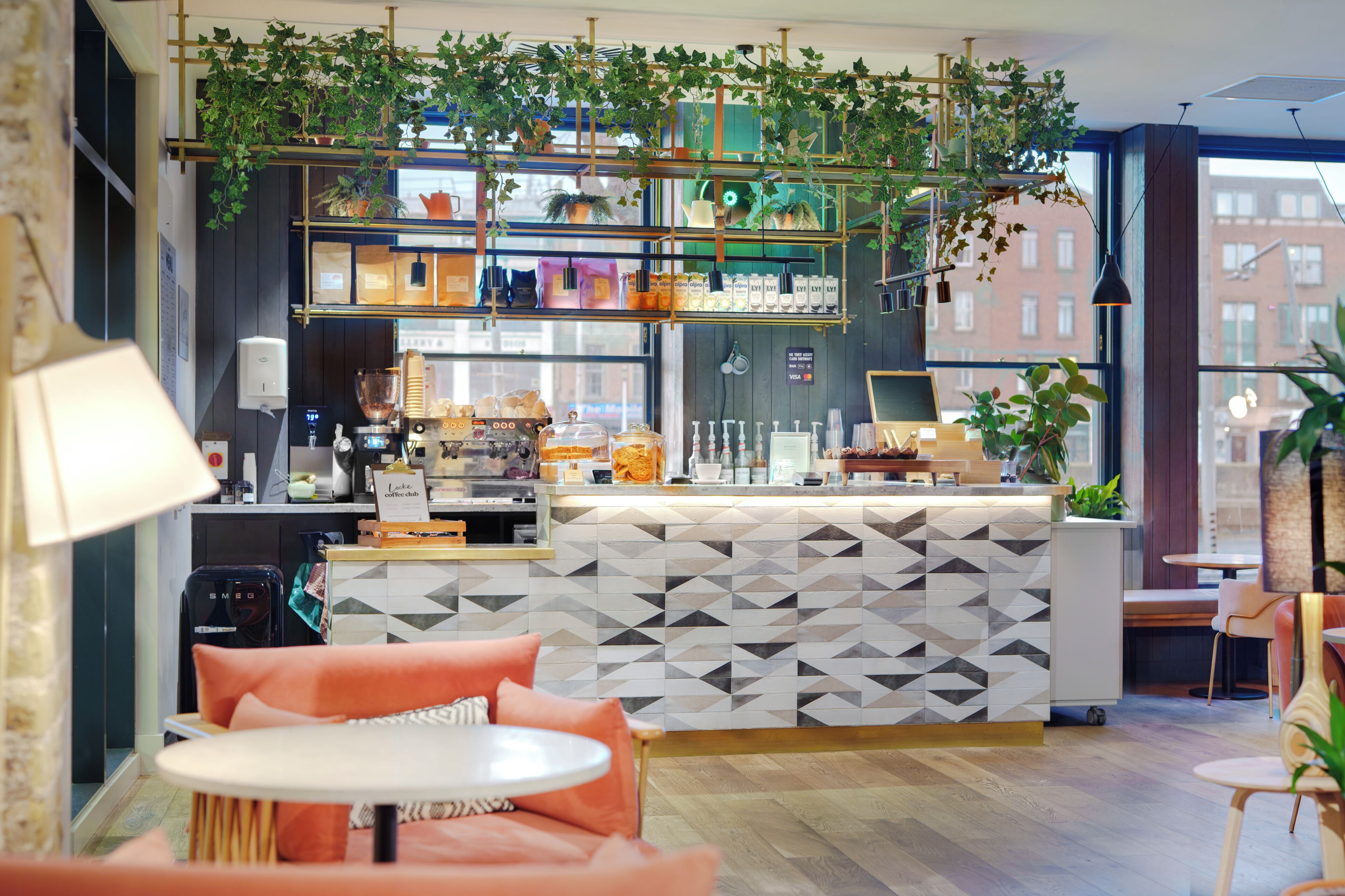 A modern cafe with a geometric patterned counter, seating with orange cushions, and hanging plants.
