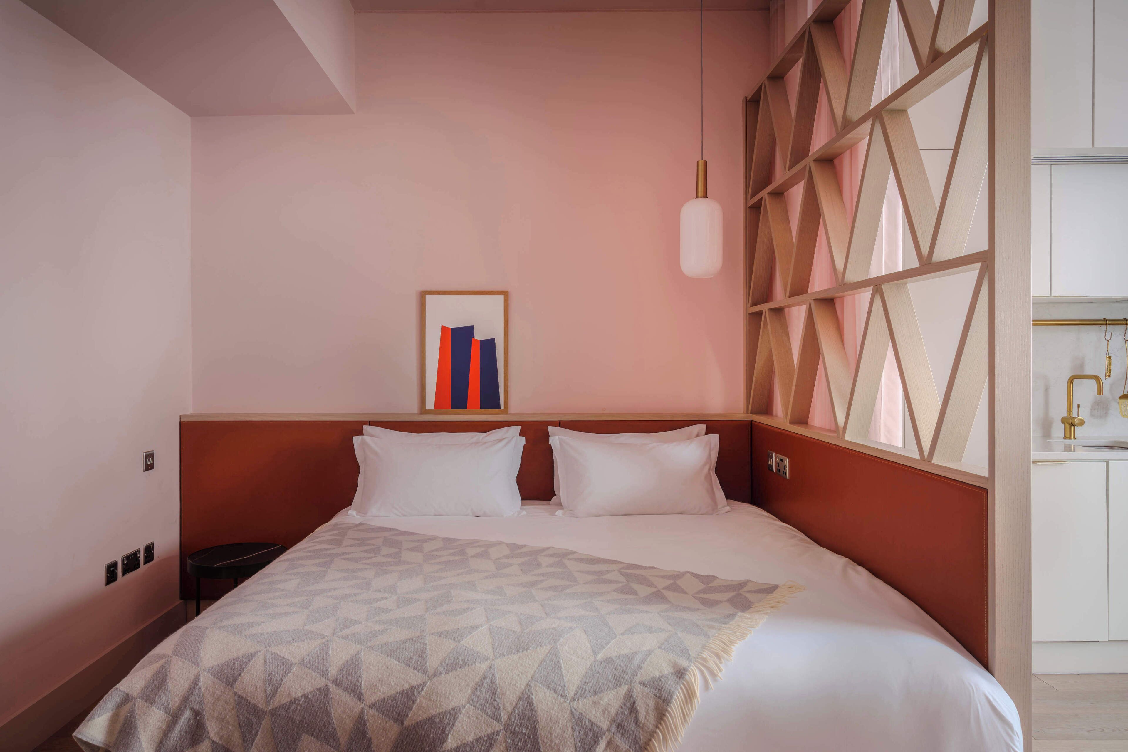 A hotel room with a bed, a headboard, a wall-mounted artwork, and a window with a geometric pattern.