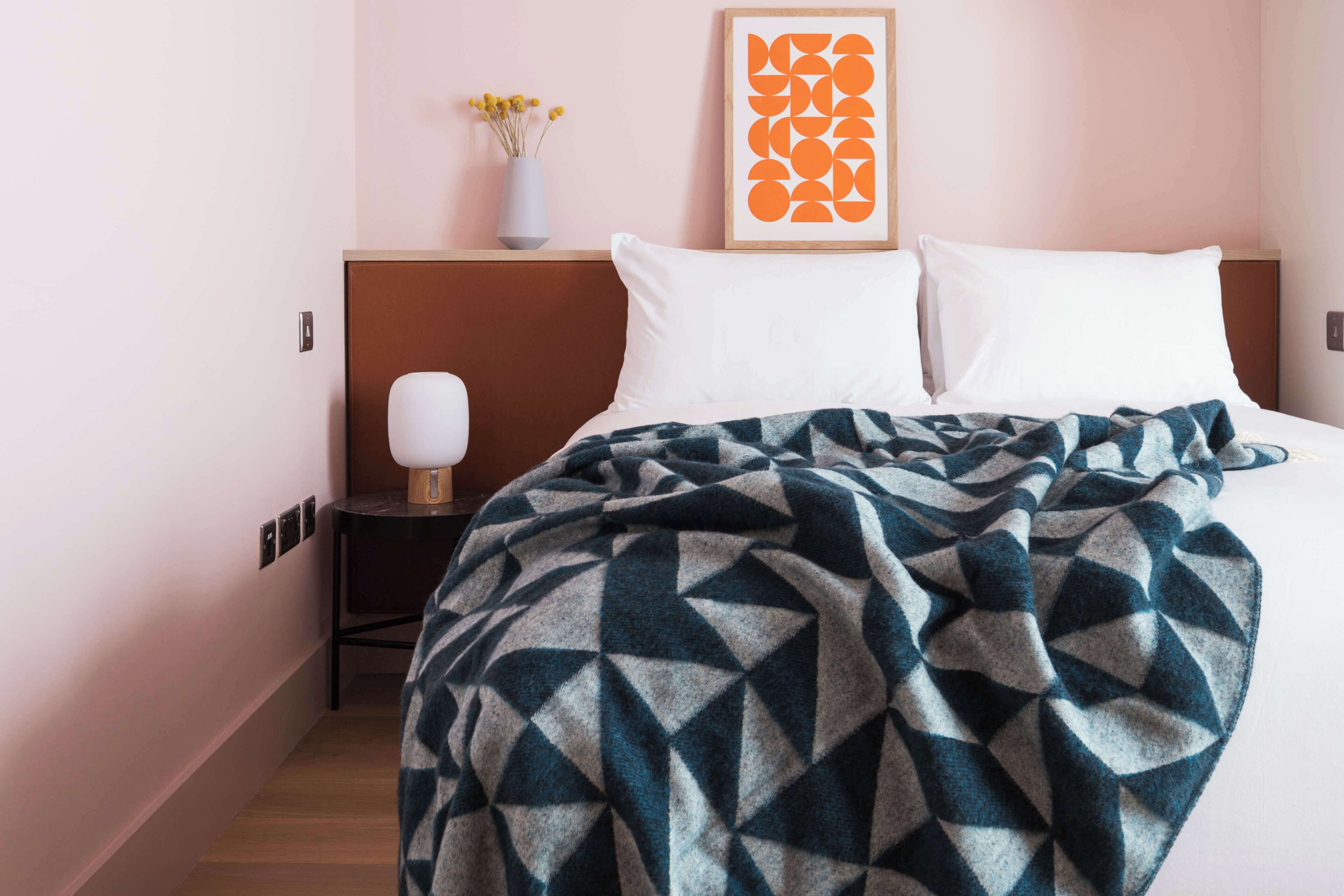A hotel room with a bed, a nightstand, a lamp, a framed artwork, and a vase with flowers.
