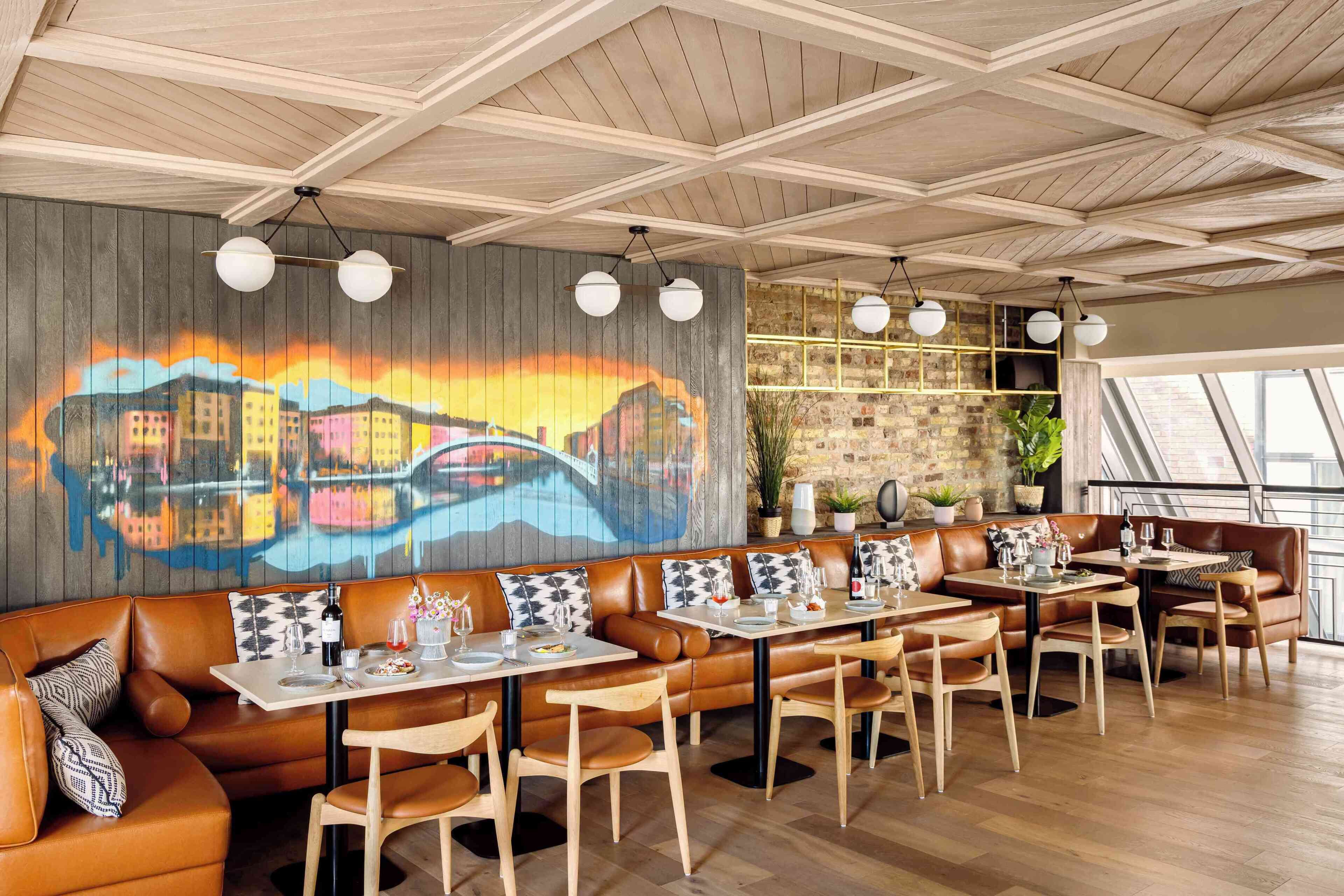 A restaurant with wooden ceiling beams, a mural, leather booths, and tables with chairs.