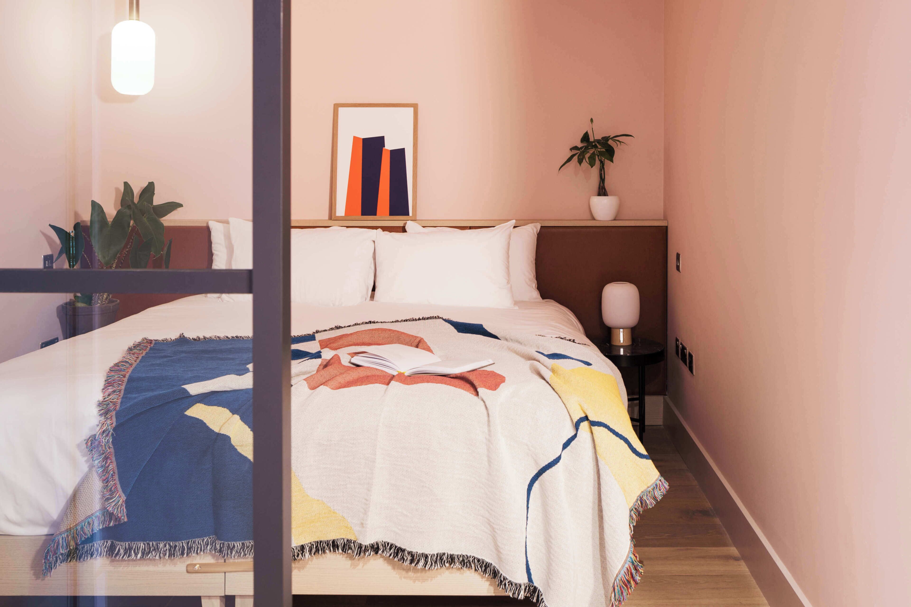 A bedroom with a bed, a bedside table with a lamp, a framed picture on the wall, and a potted plant.