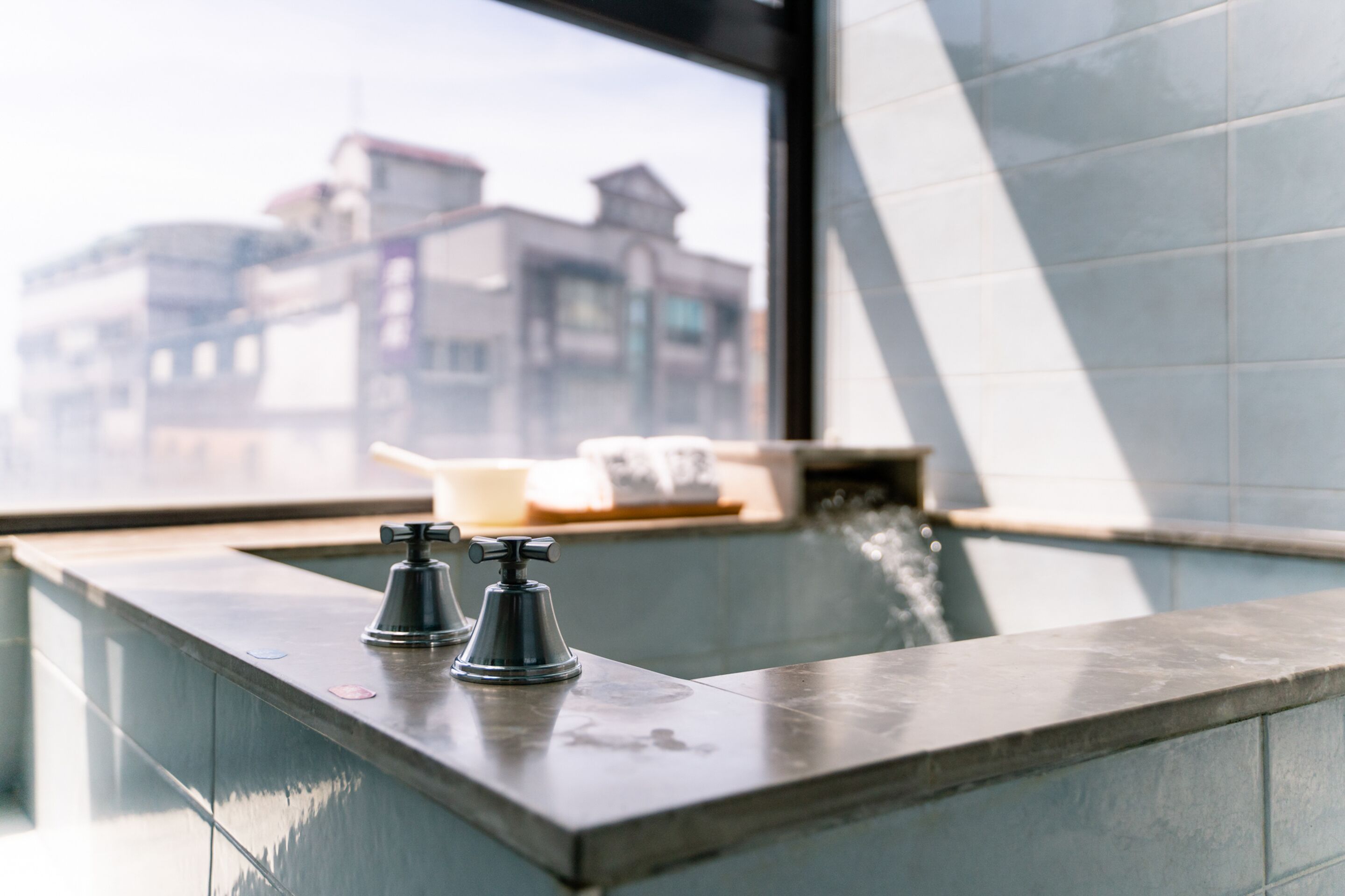 Un baño moderno con bañera, dos llaves y una ventana con vistas a edificios.