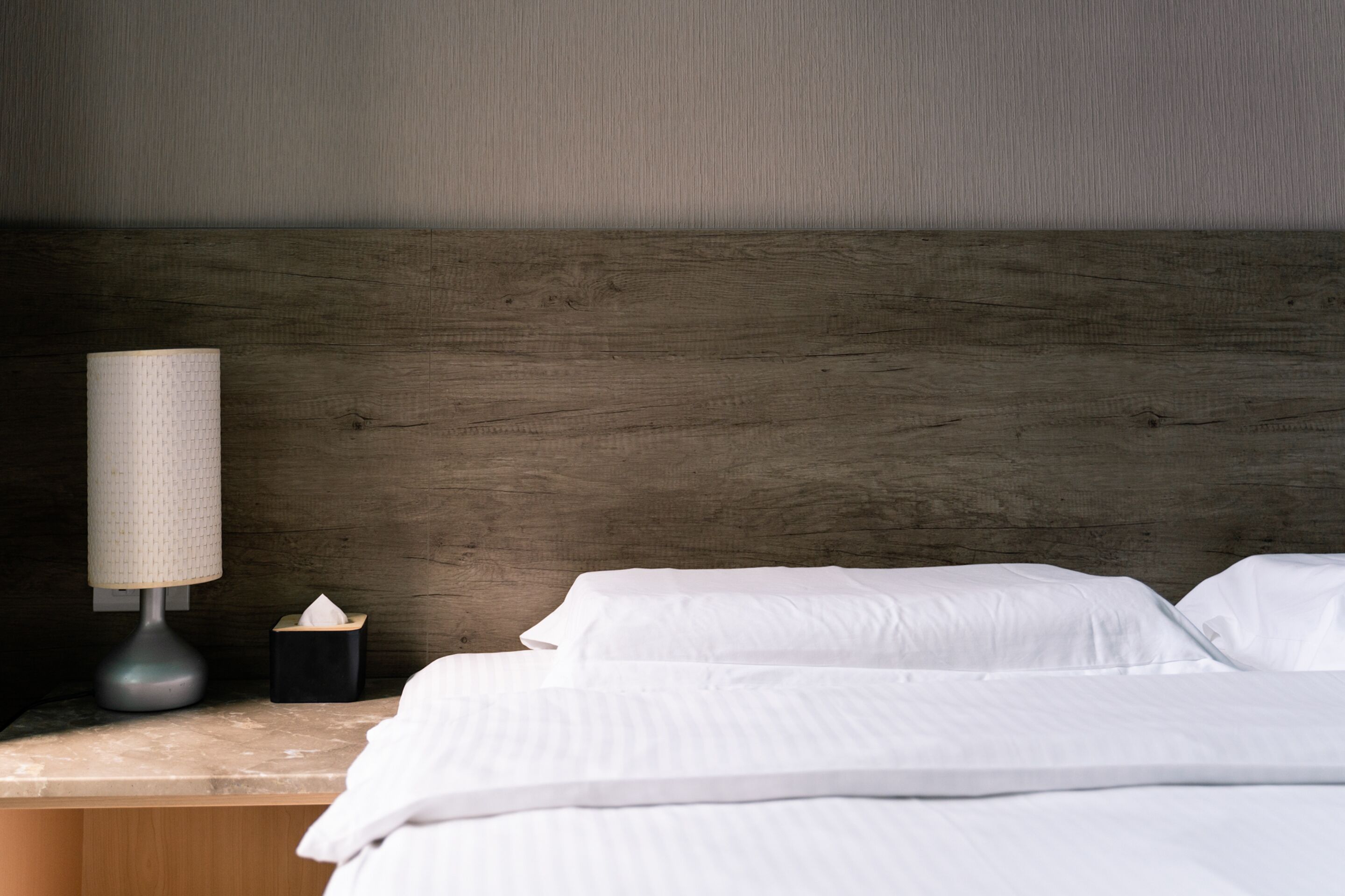 A neatly made bed with white linens, a wooden headboard, a bedside lamp, and a tissue box on a marble nightstand.