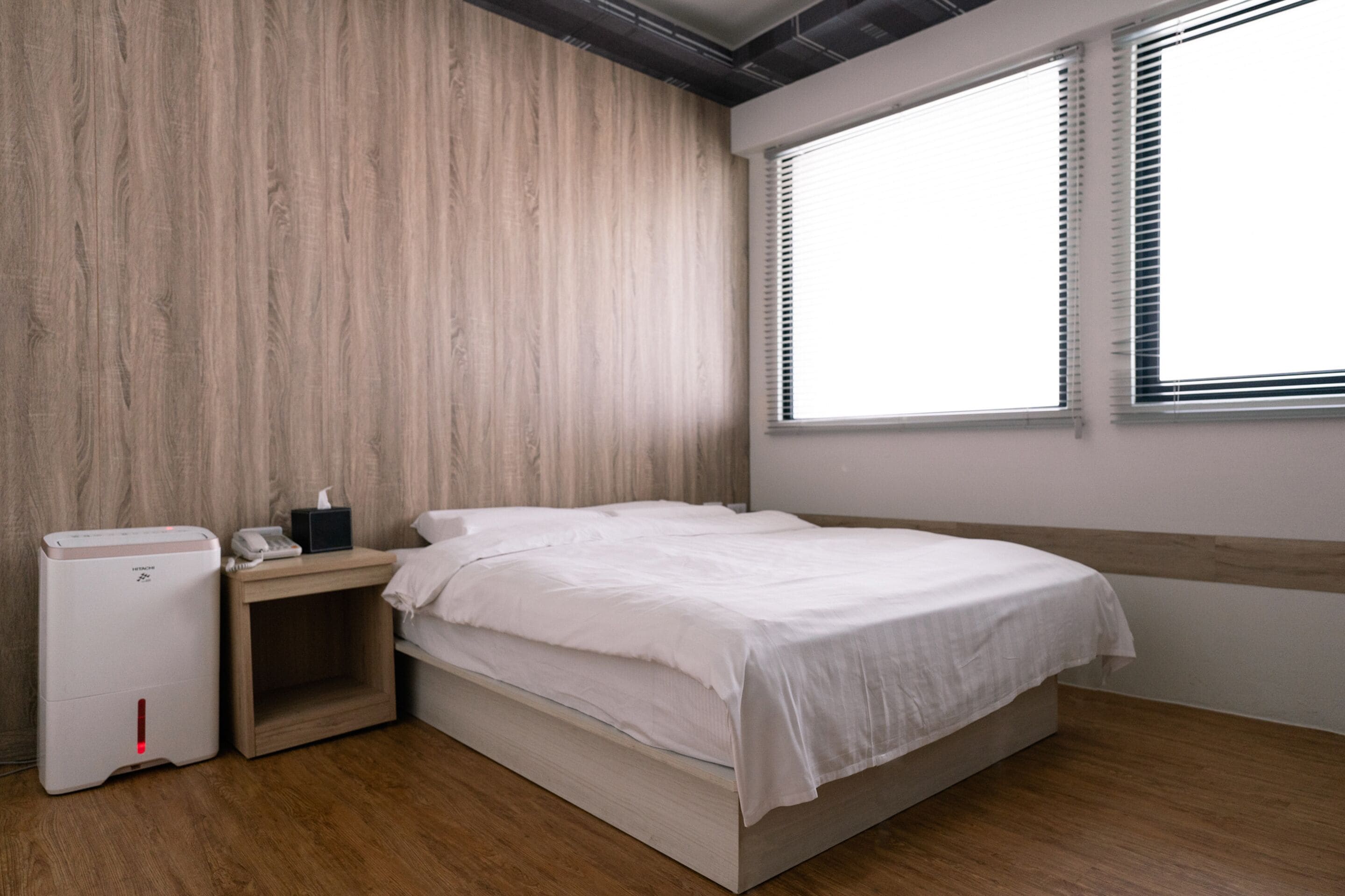 A bedroom with a wooden headboard, a bed with white bedding, a bedside table with a lamp, and a wall-mounted heater.